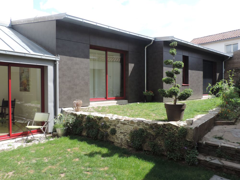 Extension à bardage sombre et grandes baies à cadres rouges ouvrant sur un jardin